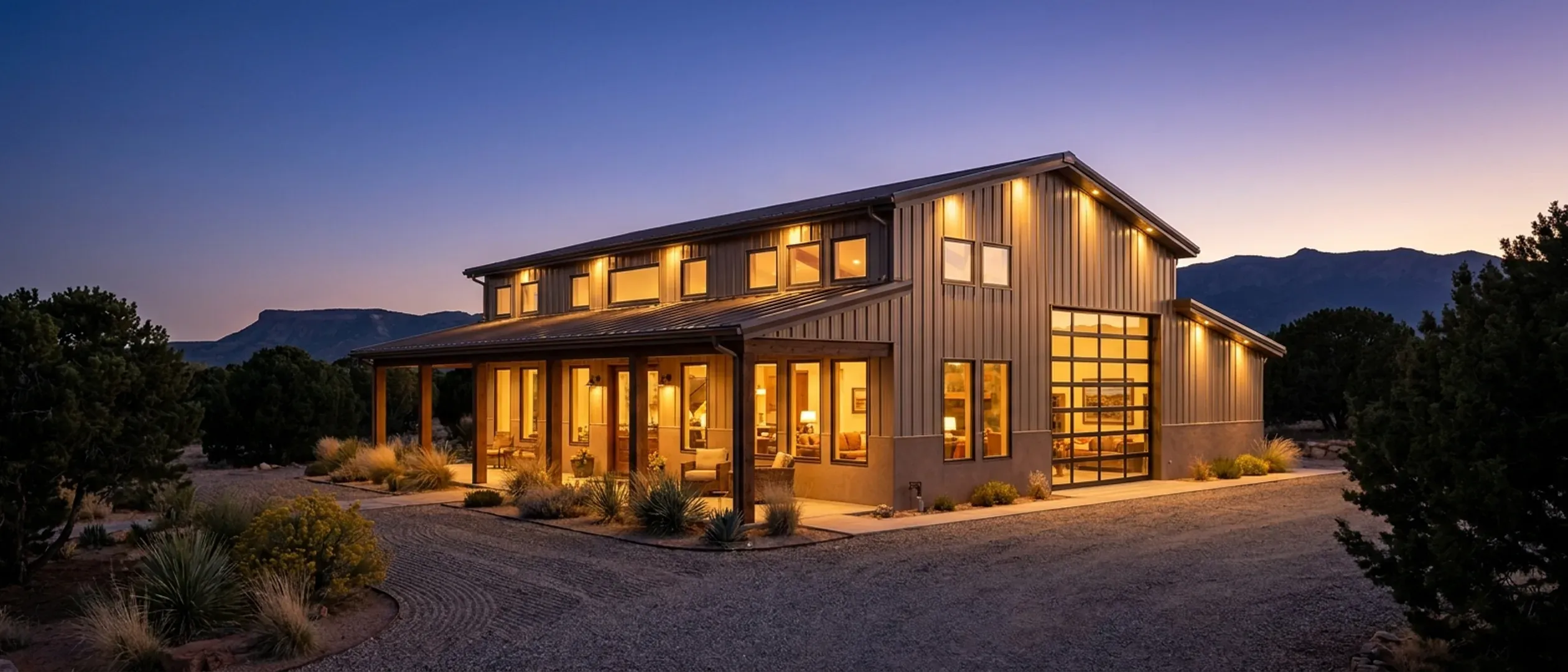 A tan metal barndominium with a deep covered porch at dusk, warm light glowing from every window, with a mesa and mountains silhouetted on the horizon.